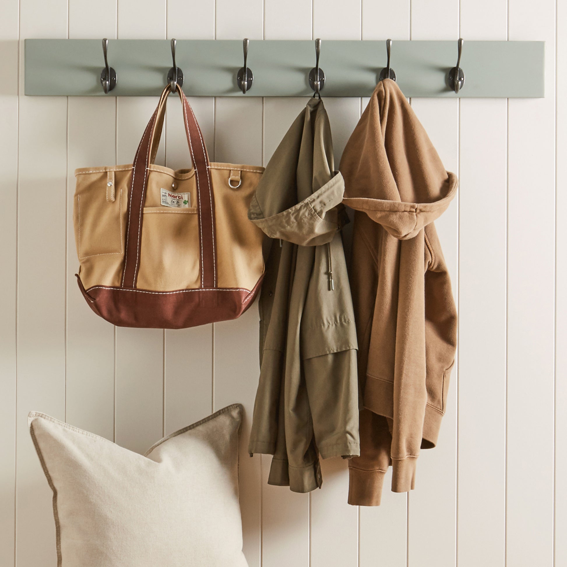 Tote bag, jackets, and a pillow hanging on a wooden hook rack against a white paneled wall.